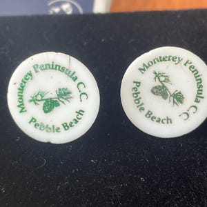 May include: Two white golf ball markers with green text and a pine tree graphic. The text reads "Monterey Peninsula C.C. Pebble Beach". The markers are circular and have a slightly worn appearance.