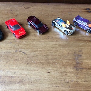 May include: Five toy cars in various colors, including black, red, burgundy, silver, and purple. The cars are arranged in a row on a wooden surface.