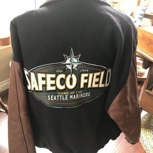 May include: A black and brown baseball jacket with a green, white, and red embroidered patch that says "Safeco Field, Home of the Seattle Mariners, Est. 1999".