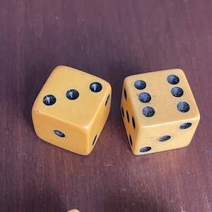 May include: Two vintage, amber-colored dice with black dots, arranged on a dark wooden surface. The dice are cube-shaped, with each side displaying a different number of dots. The dice are a classic game accessory.