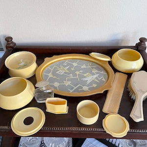 May include: A vintage vanity set with a yellow and white color scheme. The set includes a tray with a geometric design, a comb, a brush, a bottle, and several small containers.