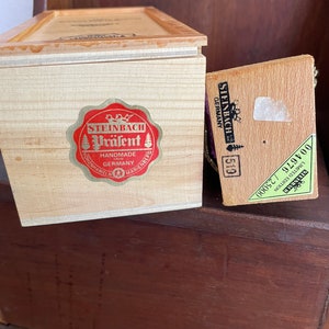 May include: Two wooden boxes with lids. The larger box has a red and white label that says "Steinbach Präzent Handmade from Germany". The smaller box has a green label that says "Steinbach Holziger Genuss 00577 929700".