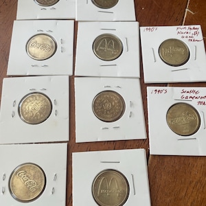 May include: A collection of vintage tokens and coins, including Coca-Cola and McDonald's branded pieces, displayed in protective white card holders. Some tokens are marked with "1990's" and location names.