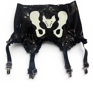 May include: A black glossy latex garter belt featuring a white pelvic bone graphic print on the front. The garment has a ruffled hemline and four adjustable black straps with silver-toned metal clips for attaching hosiery to the waistband.
