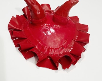 May include: A glossy red devil horn headpiece with a ruffled base. The horns are curved and pointed, and the base is circular with a pleated, layered design. The headpiece is set against a white background.