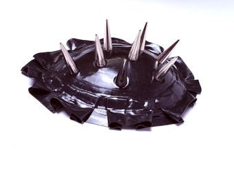 May include: A black, ruffled headpiece with multiple silver-colored spikes. The headpiece has a shiny, reflective surface and is set against a white background. The spikes are evenly spaced around the top.