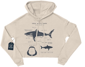 May include: A beige cropped hoodie with a graphic of a great white shark and anatomical labels. The text "GREAT WHITE SHARK" is printed on the hoodie. The hoodie has a drawstring hood.