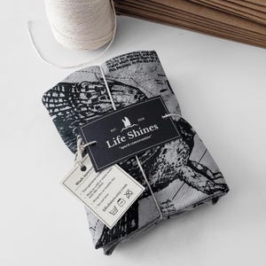 May include: A folded grey and black fabric item tied with white string, featuring a detailed illustration. The item has a tag that reads "Life Shines" and "spark conversation." A smaller tag provides washing instructions. The length is 50-66cm (20-26in).