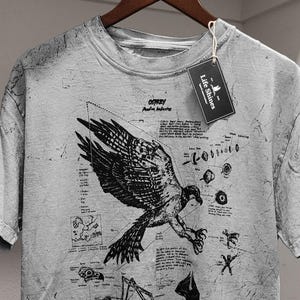 May include: A grey t-shirt featuring a detailed black illustration of an osprey with scientific diagrams and text. The shirt has a distressed appearance and a tag that reads "Life Shines".