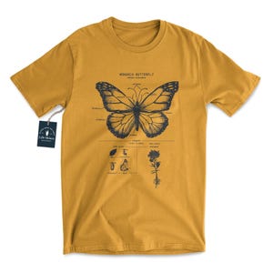 May include: A mustard yellow t-shirt with a black and white illustration of a monarch butterfly and its life cycle. The text "MONARCH BUTTERFLY" and "Danaus plexippus" is printed on the shirt.