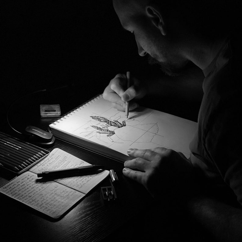 Op de afbeelding: Een persoon tekent in een schetsboek met een potlood. Het schetsboek is open op een pagina met een gedetailleerde tekening van een plant of bloem. De persoon zit aan een bureau met andere tekenmaterialen.