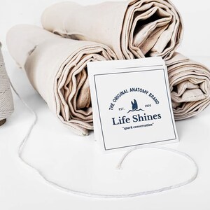 Op de afbeelding: Opgerolde witte stof met een witte kaart waarop staat "The Original Anatomy Brand Est. 2020 Life Shines "spark conversation""