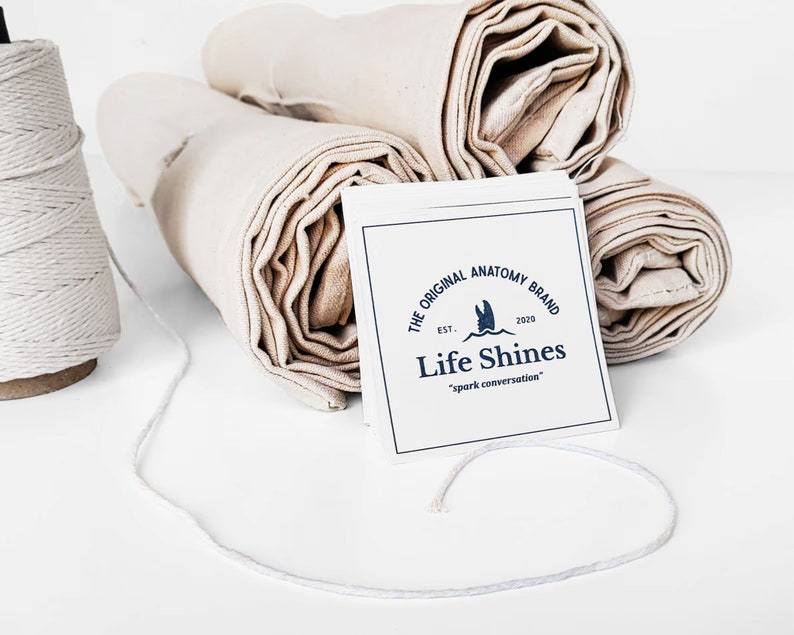 Op de afbeelding: Opgerolde witte stof met een witte kaart waarop staat "The Original Anatomy Brand Est. 2020 Life Shines "spark conversation""