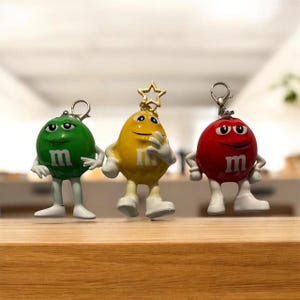 May include: Three M&M's character keychains in green, yellow, and red. Each figure has a silver keychain ring, white shoes, and the letter "m" on its front. The yellow character has a gold star on its head.