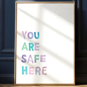 May include: A framed art print with the words "YOU ARE SAFE HERE" in colorful watercolor lettering. The print is in a gold frame and leans against a dark blue wall. The room has a wooden floor.