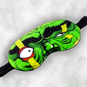 May include: A green sleep mask with a monster face design. The mask features a green face with black accents, a large white eye with a red outline, and yellow bandages. It has a black elastic strap for secure wear.