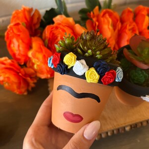 May include: A terracotta flower pot painted with a stylized face, featuring a unibrow and red lips. The pot is decorated with a colorful flower crown made of fabric.