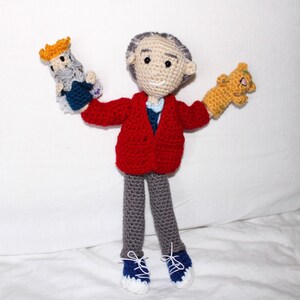 May include: A crocheted doll wearing a red cardigan, gray pants, and blue shoes. The doll is holding two smaller crocheted dolls, one with a yellow crown and the other with a brown body.