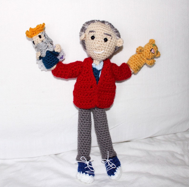 May include: A crocheted doll wearing a red cardigan, gray pants, and blue shoes. The doll is holding two smaller crocheted dolls, one with a yellow crown and the other with a brown body.