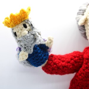 May include: A crocheted figure wearing a red jacket and holding a smaller crocheted figure wearing a yellow crown and blue and gray robes.