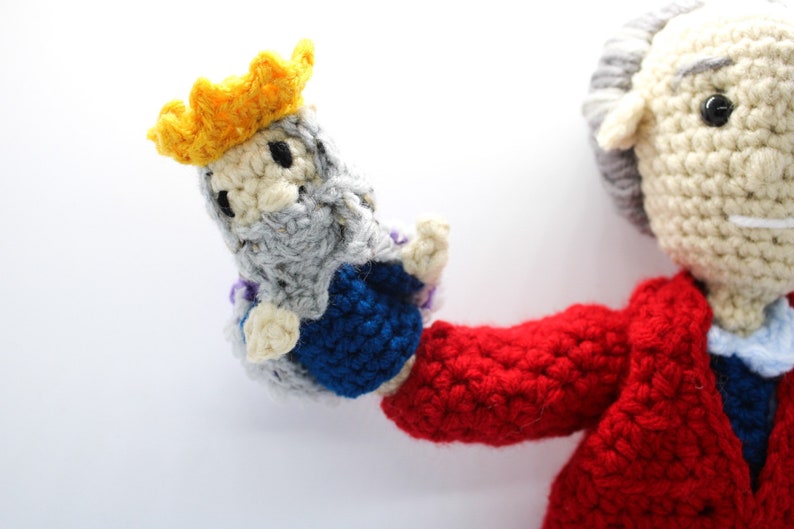 May include: A crocheted figure wearing a red jacket and holding a smaller crocheted figure wearing a yellow crown and blue and gray robes.
