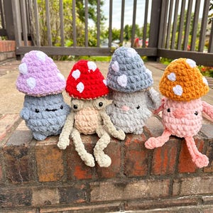 May include: Four handmade crochet mushroom plush toys in various colors: lavender, red, gray, and orange. Each mushroom has white spots and a smiling face. The toys are sitting on a brick surface.