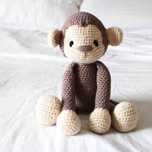 May include: A crocheted monkey toy with brown fur and beige accents. The monkey is sitting on a white bedspread.