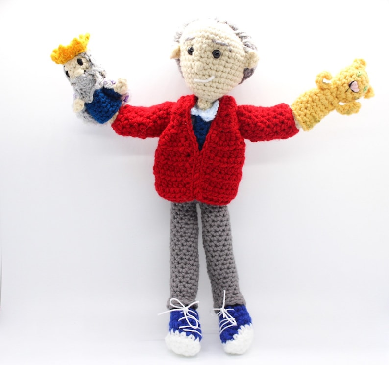 May include: A crocheted doll wearing a red cardigan, gray pants, and blue shoes. The doll is holding a crocheted puppet in each hand, one with a yellow crown and the other with orange fur.