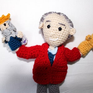 May include: A crocheted doll wearing a red jacket and gray pants, holding a small crocheted king and a crocheted cat in its hands.