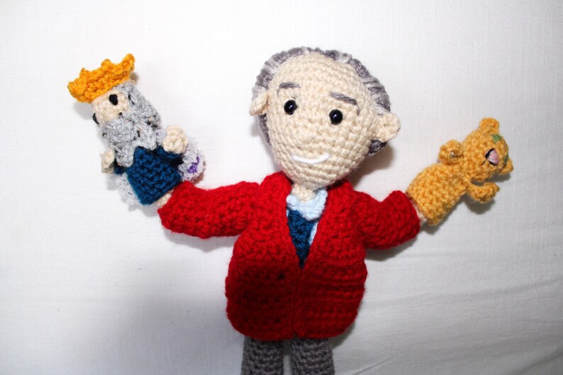 May include: A crocheted doll wearing a red jacket and gray pants, holding a small crocheted king and a crocheted cat in its hands.