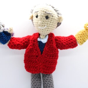 May include: A crocheted doll wearing a red jacket and gray pants, holding a crocheted puppet with a yellow crown on each hand.