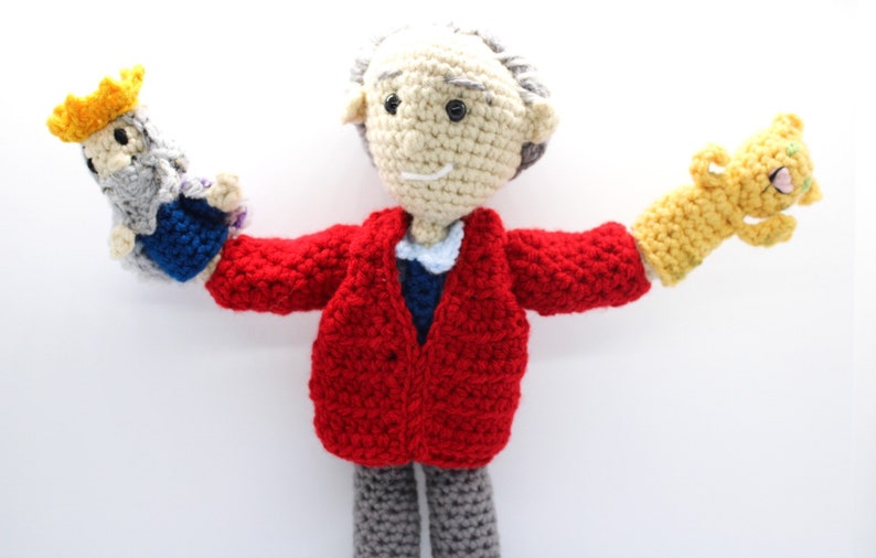 May include: A crocheted doll wearing a red jacket and gray pants, holding a crocheted puppet with a yellow crown on each hand.