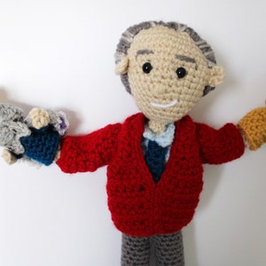 May include: A crocheted doll wearing a red sweater and gray pants, holding a gray and blue crocheted puppet in one hand and a yellow crocheted puppet in the other.