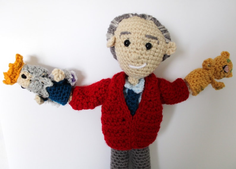 May include: A crocheted doll wearing a red sweater and gray pants, holding a gray and blue crocheted puppet in one hand and a yellow crocheted puppet in the other.