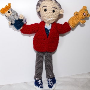 May include: A crocheted doll wearing a red jacket, gray pants, and blue shoes. The doll is holding a crocheted animal in each hand.