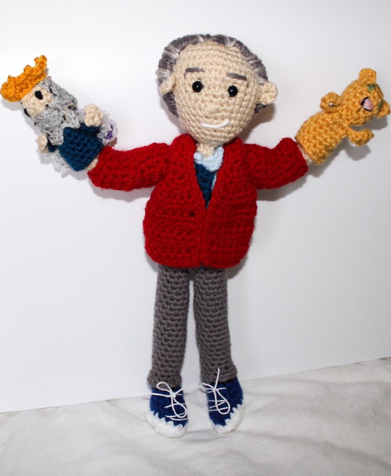 May include: A crocheted doll wearing a red jacket, gray pants, and blue shoes. The doll is holding a crocheted animal in each hand.