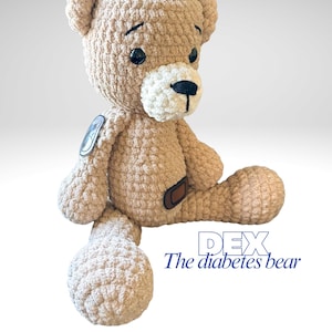 May include: A beige crocheted teddy bear with black eyes and a white muzzle. The bear has a patch on its arm and a brown patch on its leg. The text "DEX The diabetes bear" is visible.