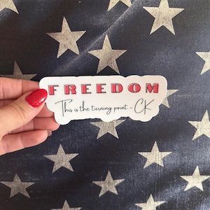 May include: A white sticker with the word "FREEDOM" in red letters. Below, the text reads "This is the turning point - CK" in a handwritten style. The sticker is held against a navy blue background with white stars.