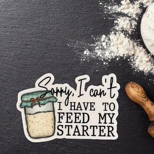 May include: A sticker with the text "Sorry, I can't, I have to feed my starter" next to a jar illustration. The jar has a green lid and a brown string tied around it. Flour and a rolling pin are in the background.