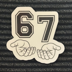 May include: A white sticker featuring the number "67" in a bold, black, collegiate-style font. Below the numbers are two stylized hands, palms up. The sticker is on a dark, textured background.