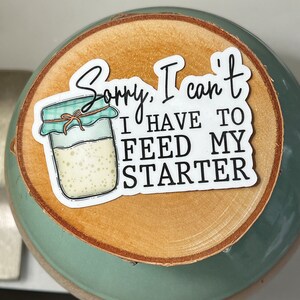 May include: A sticker with the text "Sorry, I can't, I have to feed my starter" and an illustration of a jar with a teal lid. The sticker is on a wooden slice, placed on a teal ceramic bowl.