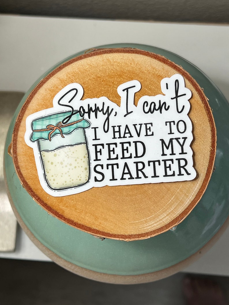 May include: A sticker with the text "Sorry, I can't, I have to feed my starter" and an illustration of a jar with a teal lid. The sticker is on a wooden slice, placed on a teal ceramic bowl.