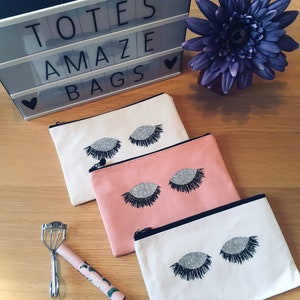 May include: Three canvas cosmetic bags in cream, pink, and white, each with a silver glitter eyelash design. A light box displays the words "TOTES AMAZE BAGS". A purple flower and a pink eyelash curler are also present.