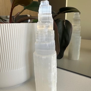 May include: A white selenite crystal tower with a layered design. The crystal is displayed on a white surface, with a white planter and a plant with dark green leaves in the background. A mirror reflects the crystal.