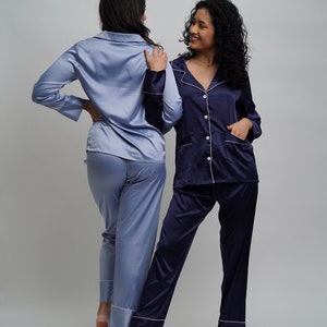 May include: Two women wearing matching satin pajamas. One woman is wearing a light blue pajama set, and the other is wearing a navy blue pajama set. Both sets have white piping around the collar, cuffs, and pockets. The women are standing facing each other and smiling.