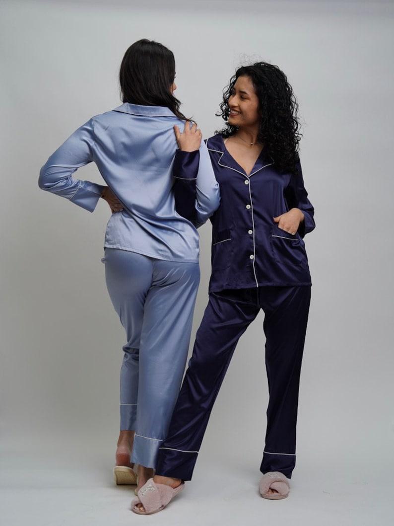 May include: Two women wearing matching satin pajamas. One woman is wearing a light blue pajama set, and the other is wearing a navy blue pajama set. Both sets have white piping around the collar, cuffs, and pockets. The women are standing facing each other and smiling.