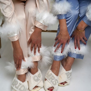 May include: Two pairs of white fuzzy slippers with the words "Bride" and "Mrs." in rhinestones. The slippers have white feather trim around the ankles.