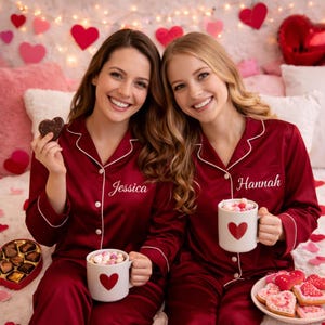 May include: Two women wearing matching burgundy pajamas with white trim, each with a name embroidered on the shirt. One holds a heart-shaped cookie, the other a mug with a heart design. The background features heart decorations.