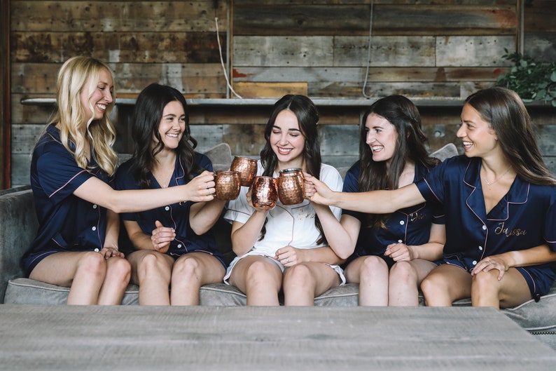 May include: Five women in navy blue pajamas with pink trim raise copper mugs in a toast. The mugs are engraved with the names "Jess" and "Dia".