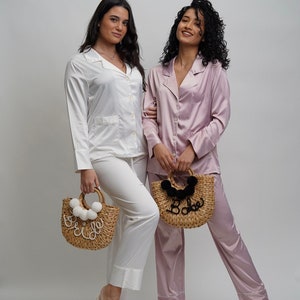 May include: Two people wearing matching satin pajamas, one white and one pink, holding straw bags with the words "Bride" and "Babe" on them. They are both wearing matching slippers with the words "Bride" and "Bridesmaid" on them.